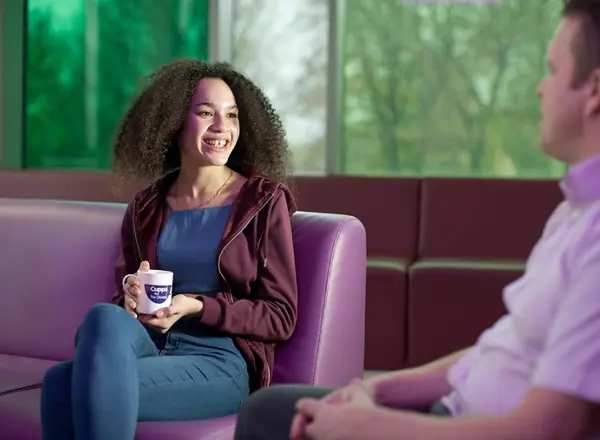 A photo of a young female Christie patient drinking a cup of tea and speaking to a man, out of shot.